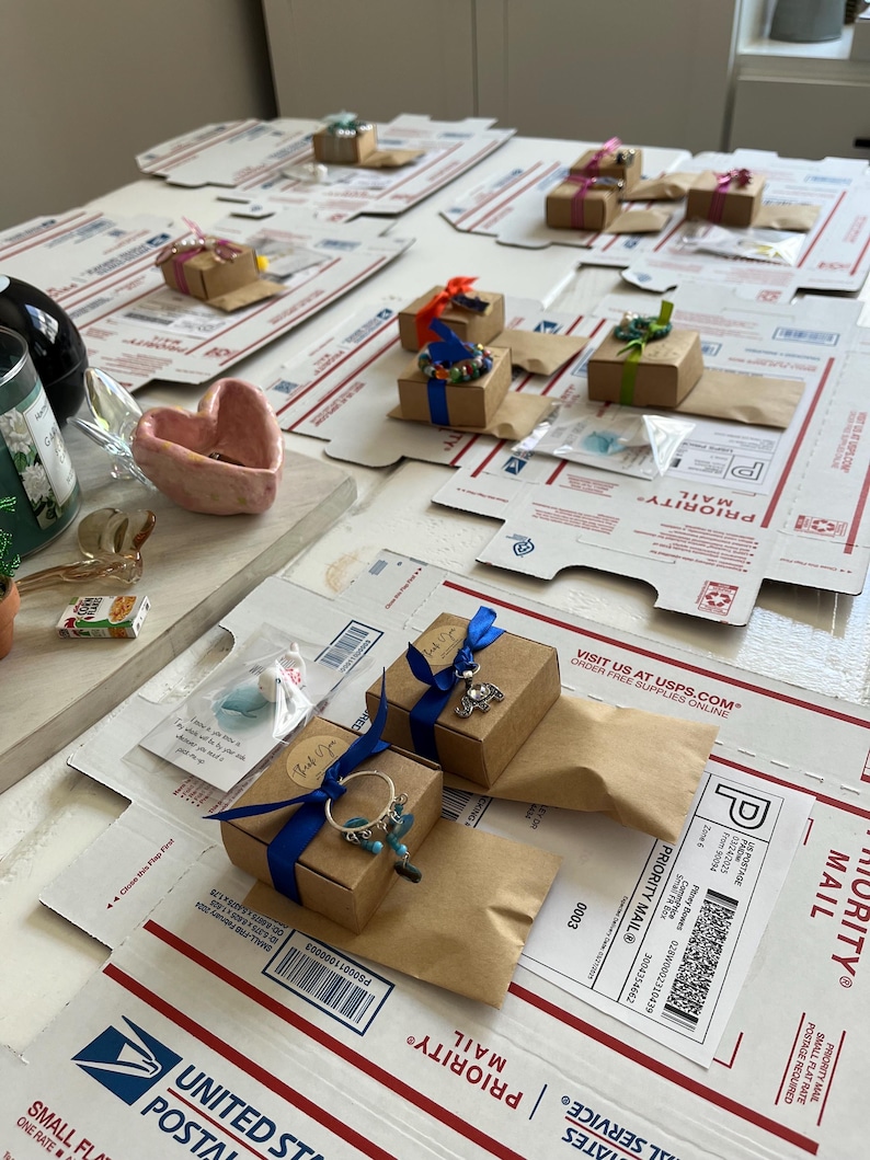 May include: A collection of small, wrapped packages containing jewelry. The packages are arranged on a white surface with brown cardboard boxes and a heart-shaped dish. The packages are tied with blue, green, and red ribbon. The packages are labeled with "PRIORITY MAIL" and "VISIT US AT USPS.COM ORDER FREE SUPPLIES ONLINE".