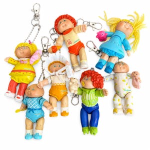 May include: A collection of seven Cabbage Patch Kids doll keychains featuring various outfits, hair colors, and accessories. Each small plastic doll is attached to a silver-toned metal lobster clasp and chain, showcasing diverse clothing styles and expressions.