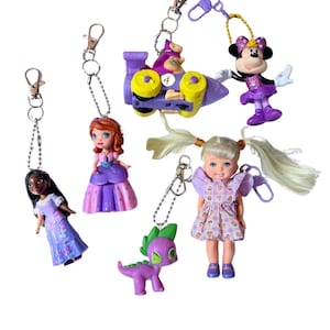May include: A collection of six character keychains featuring a doll with long blonde hair in a floral dress, a purple dragon, a princess in a purple gown, a princess in a blue and purple dress, a purple race car with yellow wheels, and a mouse in a purple dress.