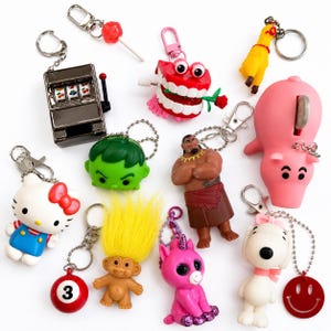 May include: A collection of assorted character keychains including a silver slot machine, chattering teeth with a rose, a yellow rubber chicken, a pink pig bank, Hello Kitty, a green Hulk head, a Maui figure, a troll, a pink unicorn, and Snoopy.