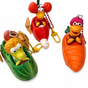 May include: Three Fraggle Rock character keychains featuring figures in vegetable-shaped vehicles. One character in a green pickle, one in a red radish, and one in an orange carrot. Each figure has a metal chain and a yellow plastic clasp attached.