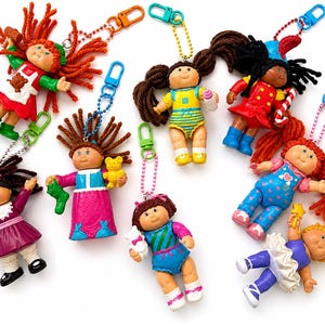 May include: A collection of seven small plastic Cabbage Patch Kids doll keychains with yarn hair, each featuring a unique outfit, colorful plastic clip, and ball chain. The dolls are depicted in various poses, some holding small accessories like toys.