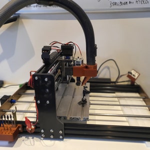 May include: A black and brown CNC machine with a vacuum hose attached to the top. The machine has a small motor and a red and brown spindle. The machine is sitting on a white surface.