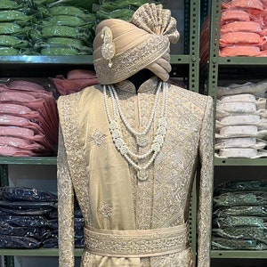 May include: A gold embroidered men's sherwani with a white pearl necklace and a gold turban. The sherwani is on a mannequin in a store.