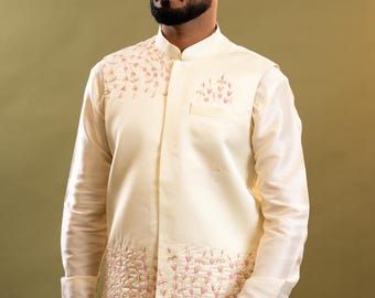 May include: Cream-colored formal wear ensemble. The outfit includes a mandarin-collared jacket with floral embroidery and a matching long-sleeved shirt and pants. The jacket features a small pocket and decorative stitching. The overall style is elegant and sophisticated.