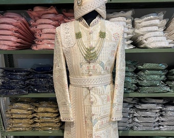 Könnte beinhalten: Ein weißer bestickter Herren-Sherwani mit einem langen, fließenden Schal und einem weißen Turban. Der Sherwani ist mit komplizierten Blumenmustern und goldenen Akzenten verziert. Der Sherwani wird auf einer Schaufensterpuppe vor einem Hintergrund aus Regalen mit gefaltetem Stoff präsentiert.