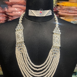 May include: A multi-strand pearl necklace with ornate, clear crystal accents. The necklace features a rectangular crystal design at the top and is displayed on a black mannequin. The brand "RICHART" is visible on a tag.