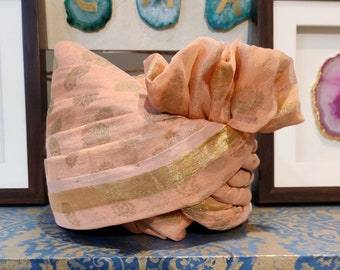 May include: A peach-coloured turban with gold trim and a ruffled top. The turban is sitting on a shelf with a blue and gold patterned surface.