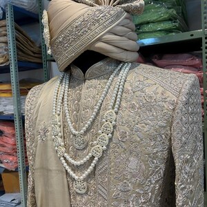 May include: A beige embroidered men's outfit with a matching turban. The outfit features intricate silver embroidery and a pearl necklace with silver pendants.