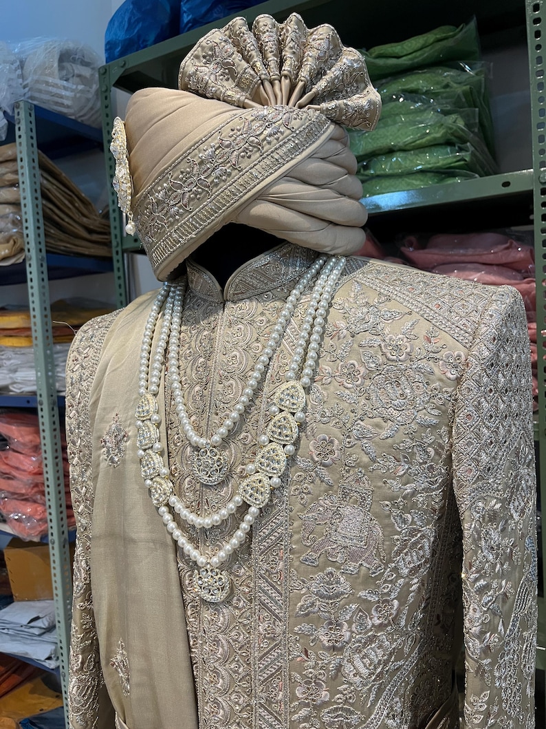 May include: A beige embroidered men's outfit with a matching turban. The outfit features intricate silver embroidery and a pearl necklace with silver pendants.