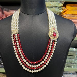 May include: A four-strand beaded necklace with white and red beads. The necklace has a gold pendant with a red gemstone.