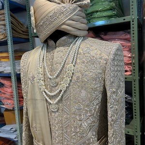 A beige embroidered men's sherwani with a silver thread design. The sherwani has a matching belt and a pearl necklace. The sherwani is worn with a beige turban with silver embroidery.