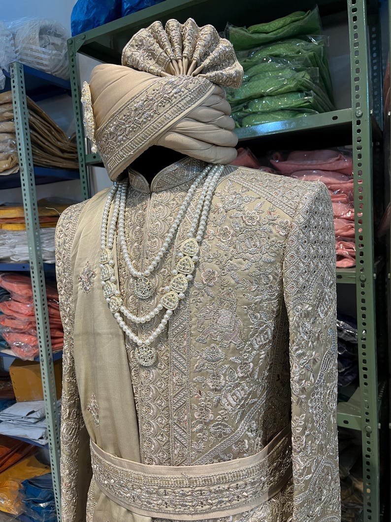 A beige embroidered men's sherwani with a silver thread design. The sherwani has a matching belt and a pearl necklace. The sherwani is worn with a beige turban with silver embroidery.