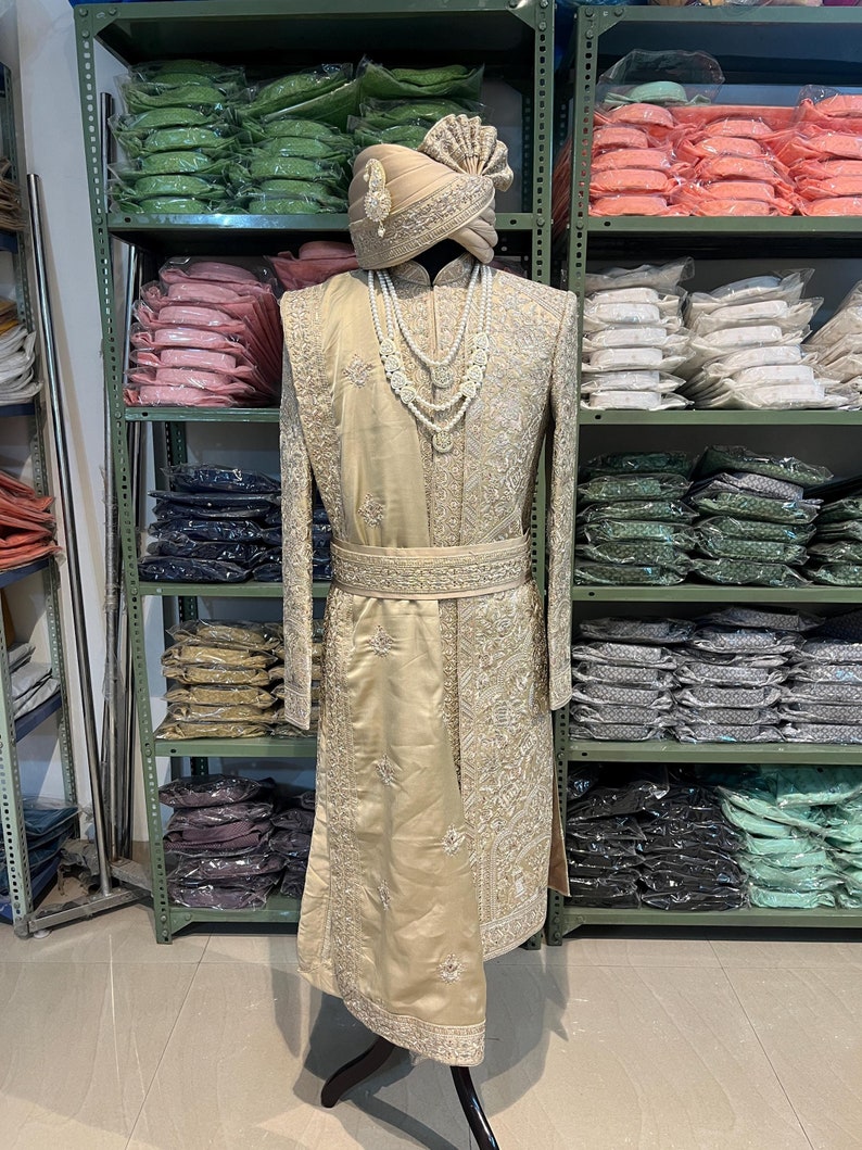 May include: A gold embroidered men's sherwani with a white pearl necklace and a gold turban. The sherwani is on a mannequin in a store.