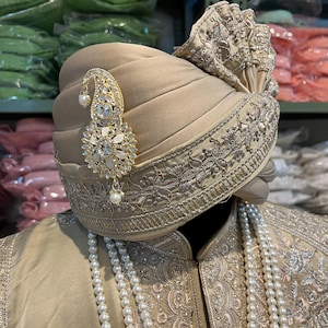 May include: A beige turban with intricate gold embroidery and a pearl and rhinestone brooch. The turban is worn over a beige outfit with a pearl necklace.