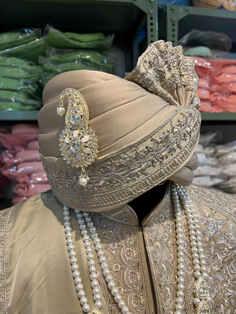 May include: A beige turban with intricate gold embroidery and a pearl and rhinestone brooch. The turban is worn over a beige outfit with a pearl necklace.