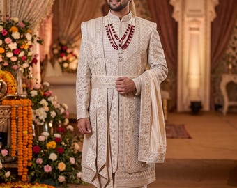 May include: A cream-coloured sherwani featuring intricate silver embroidery and a matching turban with a feather accent. The ensemble includes a layered maroon and silver beaded necklace, a draped shawl over the shoulder, and plain white trousers.