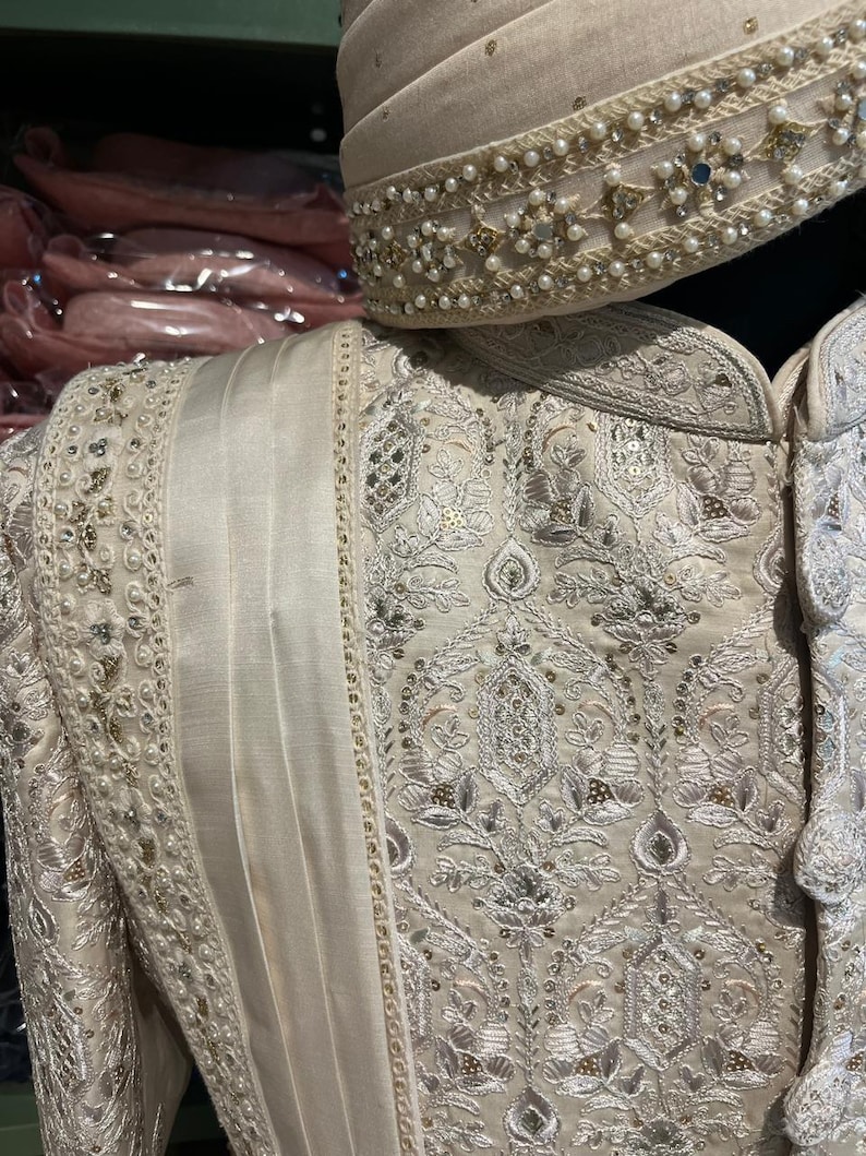 Puede incluir: Una prenda bordada de color crema con hilo plateado y detalles de perlas. La prenda tiene un cuello alto y un dise&ntilde;o largo y fluido.