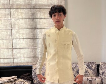 May include: A cream-colored vest with embroidered detailing, worn over a white long-sleeved shirt and white pants. The vest has a pocket on the front. The outfit is a traditional style.