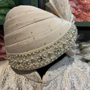 Puede incluir: Un turbante blanco con adornos de oro y perlas. El turbante est&aacute; doblado y tiene un dise&ntilde;o decorativo en el borde.