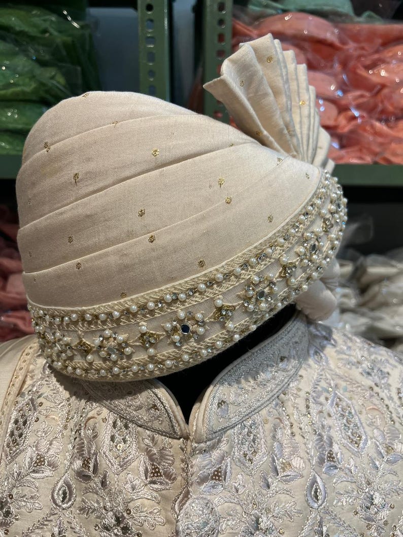 Puede incluir: Un turbante blanco con adornos de oro y perlas. El turbante est&aacute; doblado y tiene un dise&ntilde;o decorativo en el borde.