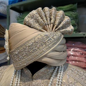 May include: A beige turban with gold embroidery and a decorative pearl and gold ornament. The turban is worn on a mannequin with a beige embroidered jacket and a pearl necklace.