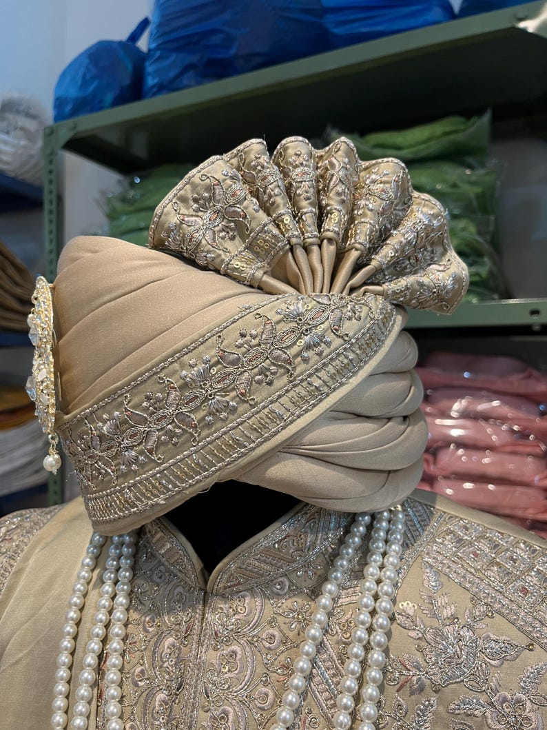 May include: A beige turban with gold embroidery and a decorative pearl and gold ornament. The turban is worn on a mannequin with a beige embroidered jacket and a pearl necklace.