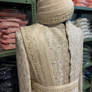 Puede incluir: Un atuendo blanco bordado para hombre con un turbante blanco a juego. El turbante tiene un ribete de cuentas. El atuendo tiene un dise&ntilde;o largo y fluido con una faja alrededor de la cintura.