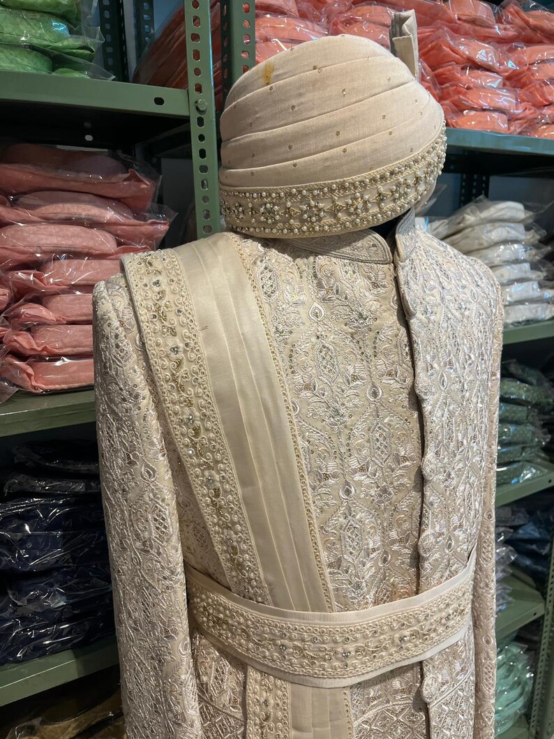 Puede incluir: Un atuendo blanco bordado para hombre con un turbante blanco a juego. El turbante tiene un ribete de cuentas. El atuendo tiene un dise&ntilde;o largo y fluido con una faja alrededor de la cintura.