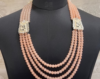 May include: A four-strand necklace made of light pink beads with gold and silver accents. The necklace has two decorative clasps with intricate designs.