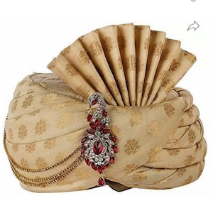 May include: A beige turban with a gold floral pattern and a decorative fan-shaped top. A jeweled brooch with red and clear stones and a gold chain adorns the turban, adding a touch of elegance. This headwear is suitable for special occasions.