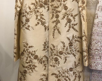 May include: Cream-coloured embroidered jacket with a mandarin collar. The jacket features intricate floral patterns in shades of brown and gold. The long-sleeved garment is displayed on a hanger, showcasing its formal design and detailed craftsmanship.
