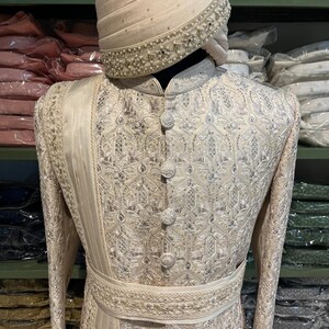 Puede incluir: Un sherwani blanco para hombre bordado con un turbante blanco a juego. El sherwani tiene un dise&ntilde;o de hilo plateado y una faja blanca. El turbante tiene un adorno de perlas.