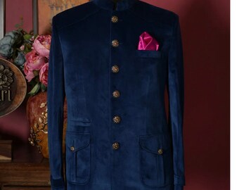 May include: A deep blue velvet bandhgala jacket featuring a mandarin collar, six ornate gold-toned buttons down the front, two lower flap pockets, and a bright magenta pocket square tucked into the left chest pocket on a mannequin torso.