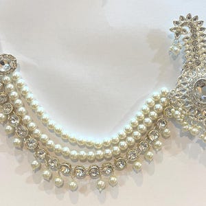 May include: An ornate, silver-toned hair accessory featuring a large, central oval-shaped clear stone, surrounded by smaller clear stones. Three strands of faux pearls cascade down, accented with more clear stones. The design includes a decorative, fan-shaped element.