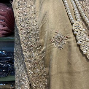 May include: A beige garment with intricate silver and pink embroidery along the edges. The garment has a pearl necklace with a large, jeweled pendant.