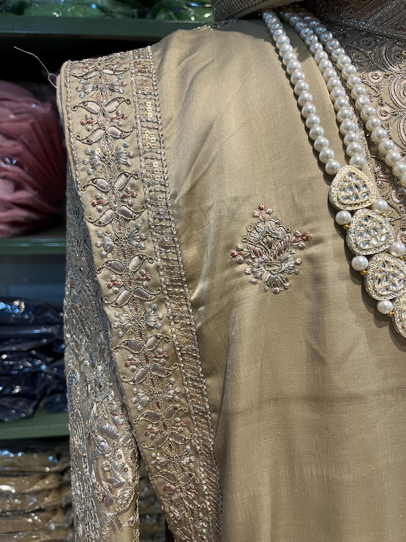 May include: A beige garment with intricate silver and pink embroidery along the edges. The garment has a pearl necklace with a large, jeweled pendant.