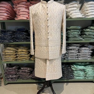 Puede incluir: Un conjunto de kurta pijama para hombre blanco bordado con cuello mandar&iacute;n y cierre de botones. El conjunto se muestra en un maniqu&iacute; sobre un fondo de estantes llenos de ropa.