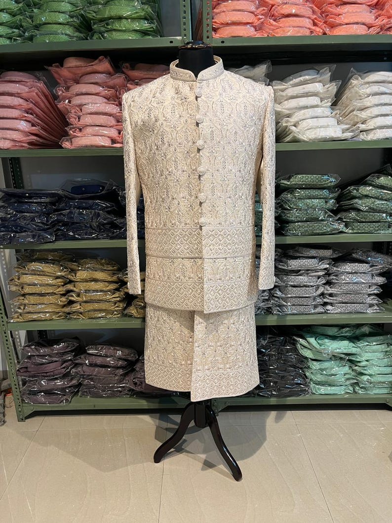 Puede incluir: Un conjunto de kurta pijama para hombre blanco bordado con cuello mandar&iacute;n y cierre de botones. El conjunto se muestra en un maniqu&iacute; sobre un fondo de estantes llenos de ropa.
