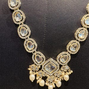 May include: A gold-toned necklace with clear, oval-shaped stones and a large, square-cut central stone. The necklace features smaller, round, clear stones around the larger stones and dangling pearls. The necklace is displayed against a black background.