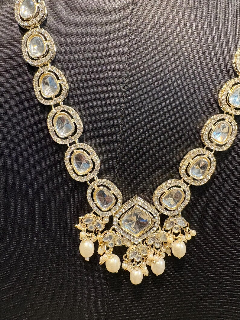 May include: A gold-toned necklace with clear, oval-shaped stones and a large, square-cut central stone. The necklace features smaller, round, clear stones around the larger stones and dangling pearls. The necklace is displayed against a black background.