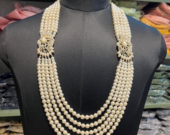 Puede incluir: Collar de perlas de cinco hilos sobre un maniquí negro. El collar presenta perlas blancas cremosas, unidas por cierres dorados adornados con cristales transparentes. Las perlas son de tamaño uniforme, creando un efecto de cascada. Una joya clásica.