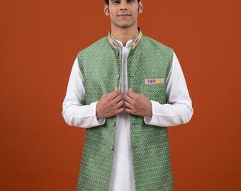 May include: A sage green sleeveless vest featuring a repeating geometric pattern, a mandarin collar with multicolored embroidery, and a matching pocket square detail. The vest is worn over a plain white long-sleeved tunic and white trousers.