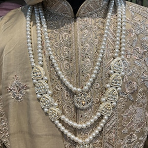 May include: A three-strand pearl necklace with decorative gold and crystal accents. The necklace is worn over a beige embroidered garment.