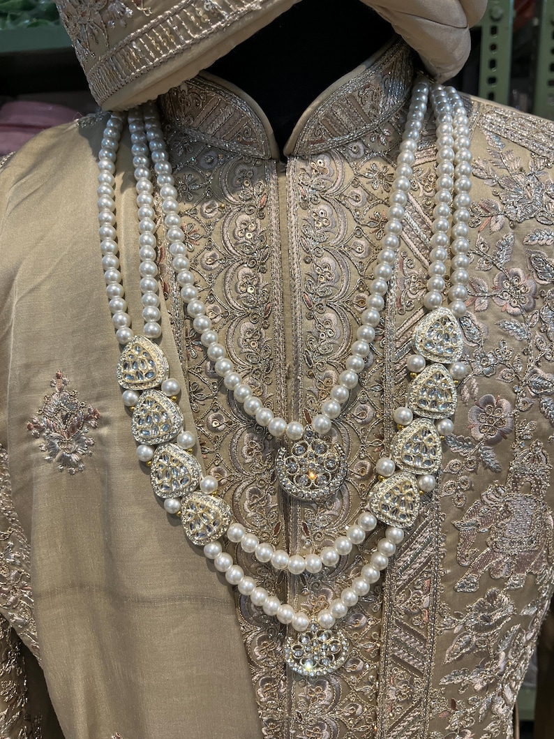 May include: A three-strand pearl necklace with decorative gold and crystal accents. The necklace is worn over a beige embroidered garment.