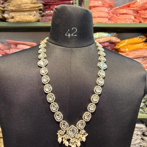 May include: A gold-toned necklace with clear, square and oval-shaped stones. The necklace is displayed on a black mannequin. The necklace has a pendant with a cluster of small, white pearls. The mannequin has the number 42 on its neck.