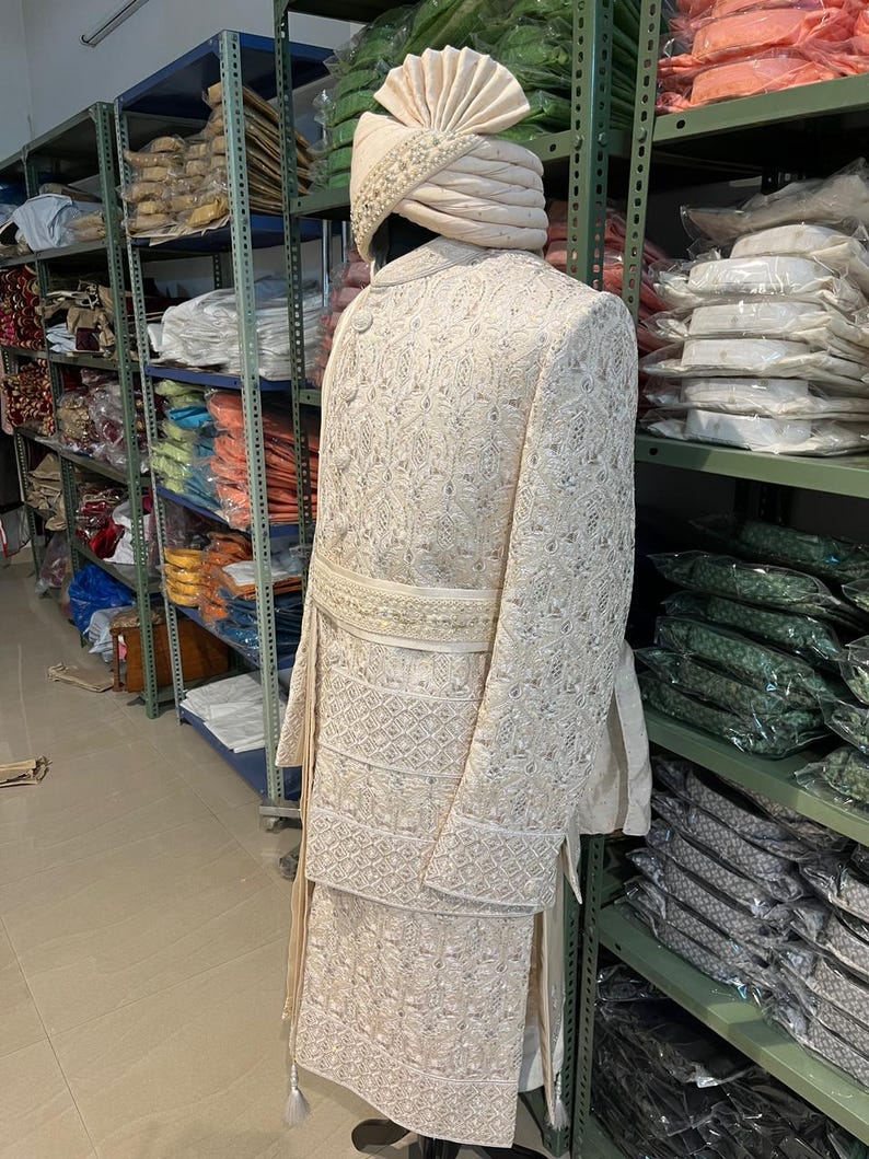Puede incluir: Un atuendo masculino blanco bordado con un turbante blanco a juego. El atuendo tiene una falda larga y fluida y una chaqueta ajustada con un cintur&oacute;n decorativo. El turbante est&aacute; decorado con perlas y una pluma blanca.