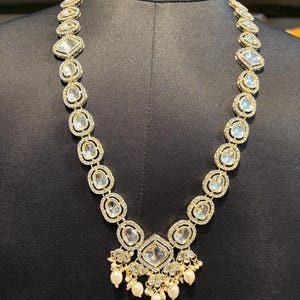 May include: A gold-toned necklace with clear, square and oval-shaped stones. The necklace features a central pendant with a large square stone and dangling pearls. The stones are set in a gold-colored metal, creating a luxurious look.
