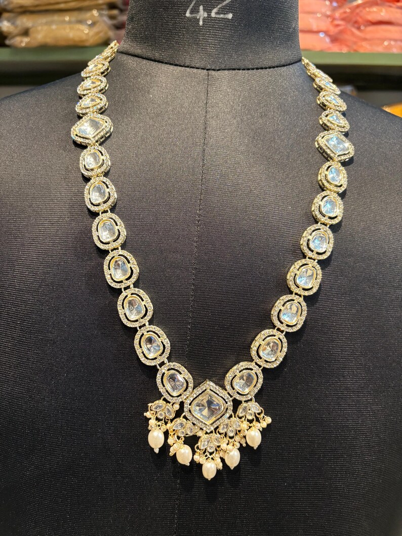 May include: A gold-toned necklace with clear, square and oval-shaped stones. The necklace features a central pendant with a large square stone and dangling pearls. The stones are set in a gold-colored metal, creating a luxurious look.
