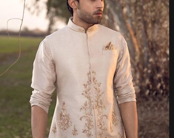 May include: A beige embroidered waistcoat with a mandarin collar and gold embroidery. The waistcoat has a pocket on the left side with a gold patterned handkerchief.